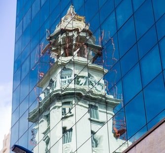 Reflection of a historic white clock tower with intricate details on the glass facade of a modern blue building in Uruguay. The tower's image is slightly distorted due to the glass's reflective properties, much like an entity health check revealing underlying nuances.