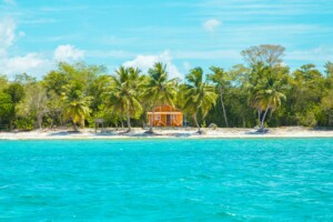 A small, orange wooden house with a white fence sits on a sandy beach surrounded by palm trees and lush greenery. The beach faces turquoise blue water under a sunny sky with light clouds, providing an idyllic backdrop reminiscent of where you might find a lawyer in the Dominican Republic relaxing.