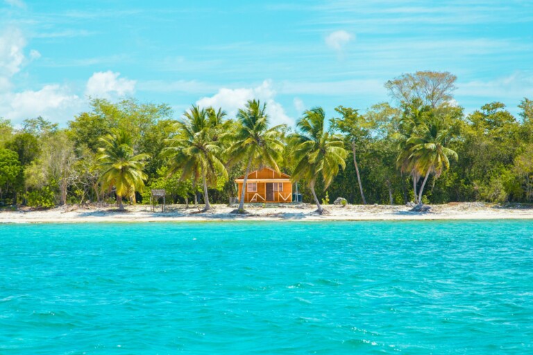 A small, orange wooden house with a white fence sits on a sandy beach surrounded by palm trees and lush greenery. The beach faces turquoise blue water under a sunny sky with light clouds, providing an idyllic backdrop reminiscent of where you might find a lawyer in the Dominican Republic relaxing.