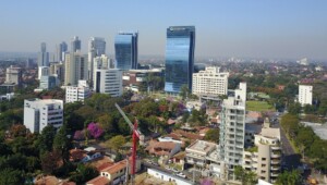 Aerial view of a cityscape featuring a mix of high-rise buildings and smaller structures. The skyline, perhaps observed by a lawyer in Paraguay, includes several modern glass towers and ongoing construction. Trees and greenery are interspersed among the buildings, with a clear sky overhead.