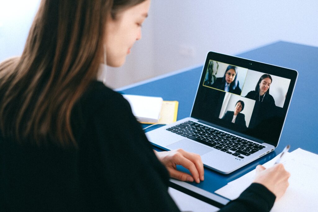 A person is having a virtual meeting using a laptop. The screen displays four individuals in a video conference discussing Bolivia tech companies. The person appears to be taking notes with a pen and paper, positioned on a blue table.