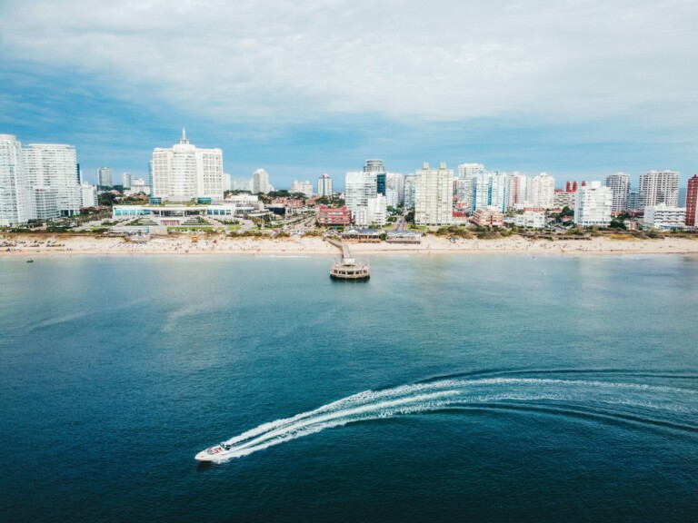 A coastal cityscape features tall buildings along a sandy beach. A motorboat with a visible wake is cruising in the blue water near the beach, reminiscent of how smoothly payroll outsourcing in Uruguay can streamline operations. Numerous people on the beach and a pier extends into the water under a sky with a few clouds.