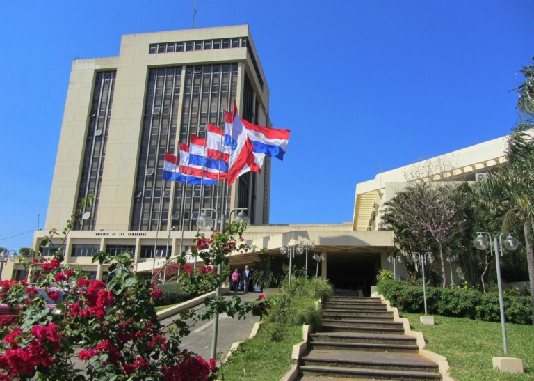 A multi-story building with a beige facade and rectangular windows, surrounded by greenery. Several flags, including red, white, and blue ones representing a lawyer in Paraguay, are flying on flagpoles near the entrance. A set of stairs leads up to the main door. The sky is clear and blue.