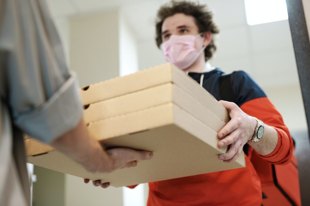 A person wearing a pink mask, red and blue jacket, and a wristwatch is handing over three pizza boxes to another individual indoors, with a backdrop of off-white walls and a ceiling light. The setting could easily be mistaken for break time at one of Ecuador's tech companies.