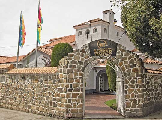 A stone-walled building with a gated archway displays a sign reading “Autoridad de Impugnación Tributaria” in Spanish, marking the fiscal address in Bolivia. Two flags stand on flagpoles near the white exterior and red-tiled roof. Nearby, trees add a touch of greenery to the scene.