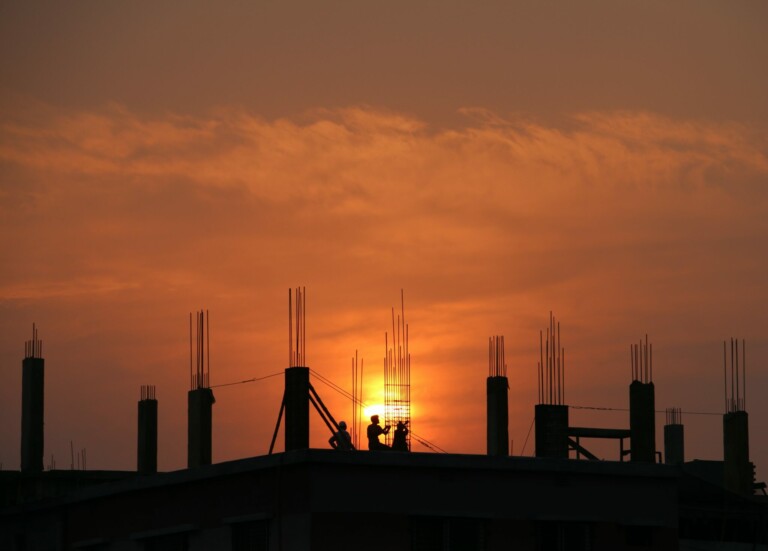 Silhouetted against an orange sunset, several workers from Construção Civil do Brasil are seen on the rooftop of a building under construction. Reinforced columns with exposed rebar protrude from the structure, and the sky is filled with warm, gradient hues.