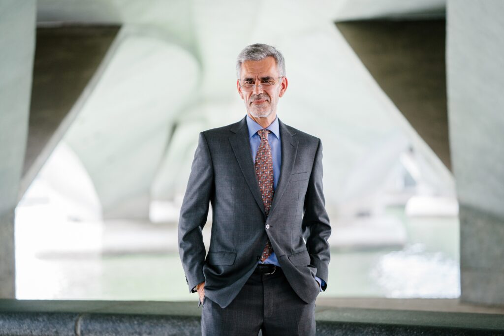 A local director in Argentina with gray hair and a beard stands under a concrete structure with geometric designs. He is wearing a dark gray suit, a light blue shirt, and a red patterned tie. With his hands in his pockets, he looks directly at the camera.