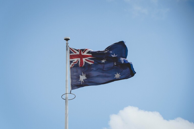 An Australian flag is flying on a flagpole against a backdrop of a blue sky with a few clouds, symbolizing the optimism and potential for starting a business in Australia. The flag features the Union Jack in the upper left corner and several white stars on a dark blue field.