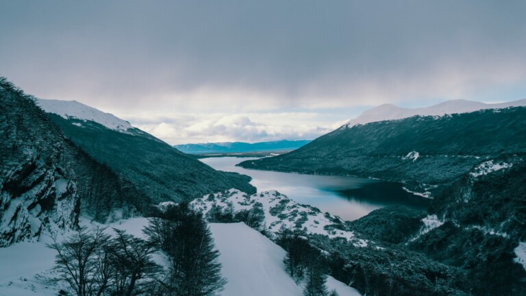 A serene winter landscape featuring a snow-covered mountain valley with a narrow body of water flowing through it, reminiscent of scenes captured by a local director in Argentina. The sky is overcast, and the mountains in the distance are also blanketed in snow. Bare trees populate the foreground.