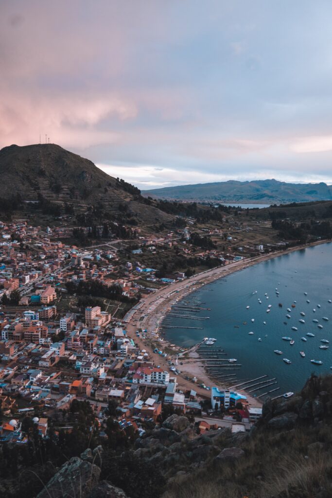 A coastal town with numerous buildings sits next to a curved shoreline. Boats are docked in the water along the edge. Hills and mountains, reminiscent of Bolivia's terrain, are seen in the background under a cloudy sky during twilight, while tech companies drive innovation locally.