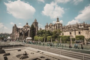 The image shows a street view with people walking, featuring a historic cathedral in the background under a partly cloudy sky. The foreground includes a glass barrier and what appears to be an archaeological site or modern construction elements, possibly blending old charm with the bustling life of a lawyer in Mexico.