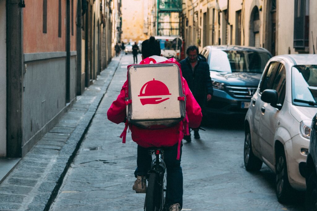 A delivery person wearing a red jacket and black beanie rides a bicycle down a narrow street. They have a large, square delivery bag with a food logo on their back, reminiscent of the innovative spirit seen in Paraguay startups. The street is lined with buildings, and a few pedestrians and parked cars are visible.