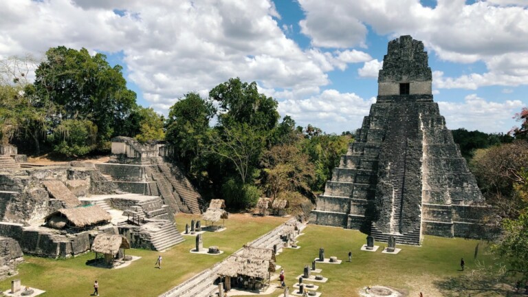 A wide-angle view of ancient Mayan ruins, with a tall stone pyramid standing prominently on the right. Various smaller structures surround the area, set against a backdrop of green trees and a partly cloudy sky. Visitors are walking among the ruins, contemplating buying property in Guatemala nearby.