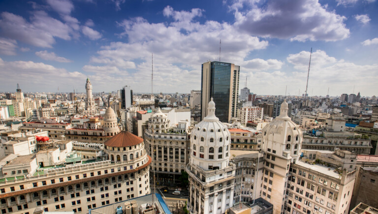 Aerial view of a cityscape featuring a mix of historical and modern architecture. Prominent buildings include ornate, dome-topped structures in the foreground, and a tall, modern skyscraper in the center. The sky is partly cloudy, with multiple tall antennas visible, reflecting the bustling hub for legal services in Argentina.