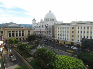 A wide-angle view of a city square with a mix of historic and modern buildings. The center features a park with trees and a fountain. Prominent structures include a white domed building in the background and, to the right, a multi-story yellow building that houses the most renowned lawyer in El Salvador.