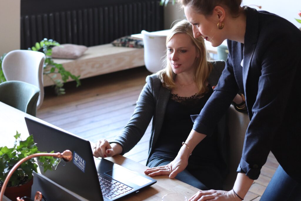 Two women are working together in an office space. One woman is seated, looking at a laptop, while the other stands beside her, pointing at the screen. The workspace, ideal for M&A Due Diligence in Colombia, includes modern furniture and some green plants.