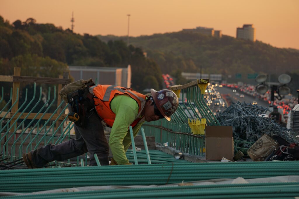 A construction worker wearing a safety vest and helmet bends down to adjust materials on a bridge under construction at sunset. The worker is surrounded by equipment, while in the background, a busy highway with cars and distant hills set the scene, reminiscent of El Salvador tech companies' bustling environments.