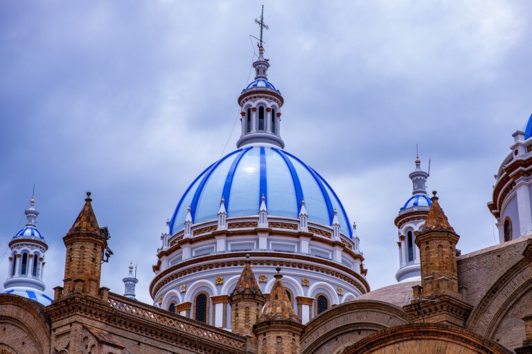 Una imagen detallada de la cúpula de una catedral con franjas azules y blancas, rodeada de cúpulas más pequeñas y torreones de ladrillo marrón, evoca el encanto de Ecuador. El cielo está nublado.