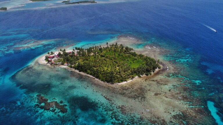 Aerial view of a small, lush island surrounded by clear blue waters and coral reefs. There are some buildings among the trees, perhaps housing Corporate Legal Services in Panama, and a boat sailing in the distance. The island is encircled by shallow, turquoise waters that transition to deeper blue.