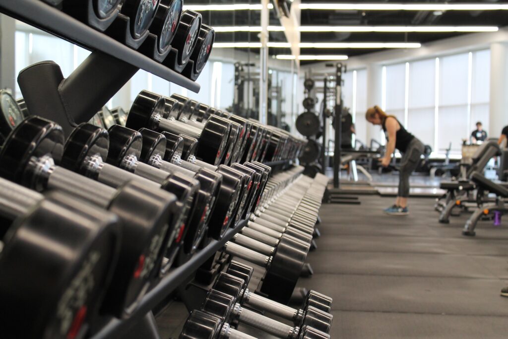 A gym interior with rows of dumbbells neatly arranged on a rack in the foreground. In the background, a person wearing workout attire is bending over, possibly picking up weights. Various other gym equipment and users, some discussing Paraguay startups, are visible further in the distance.