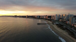 A coastal cityscape during sunset with a series of high-rise buildings lining the shore. The beach stretches along the right side of the image while the calm sea extends to the left. An office for an advogado corporativo no Uruguai stands out among the skyline. The sky is partly cloudy with hues of orange and purple.