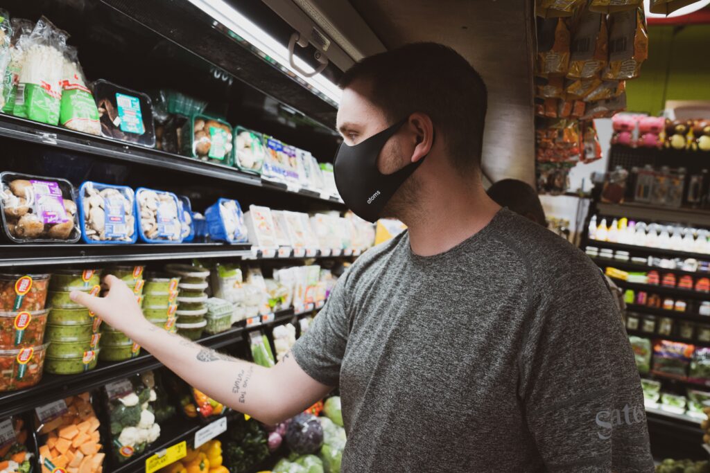 A man wearing a black face mask and a gray shirt is standing in a grocery store, reaching for a container of food on a refrigerated shelf. The shelves are stocked with various packaged vegetables and other items, reflecting the diverse range typical of Paraguay startups. The store lighting is bright.