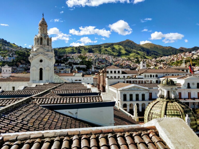 A scenic view of a historic city with a foreground of rooftops featuring clay tiles. The city, perfect for product registration in Ecuador, includes prominent white buildings with ornate architecture and a large dome. Hills covered with greenery and scattered houses form the background under a clear blue sky.