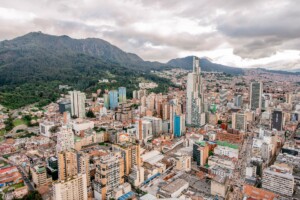 Aerial view of a cityscape featuring numerous high-rise buildings and densely packed structures. In the background, a green mountain range is visible under a cloudy sky. The city, possibly home to a thriving lawyer in Colombia, appears to be nestled in a valley.