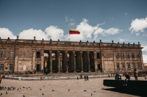 A historical building with columns and a Colombian flag on the roof stands under a blue sky with scattered clouds. The courtyard in front has a few people walking, and there are pigeons on the ground, creating an atmosphere where M&A due diligence in Colombia might feel both serene and diligent.