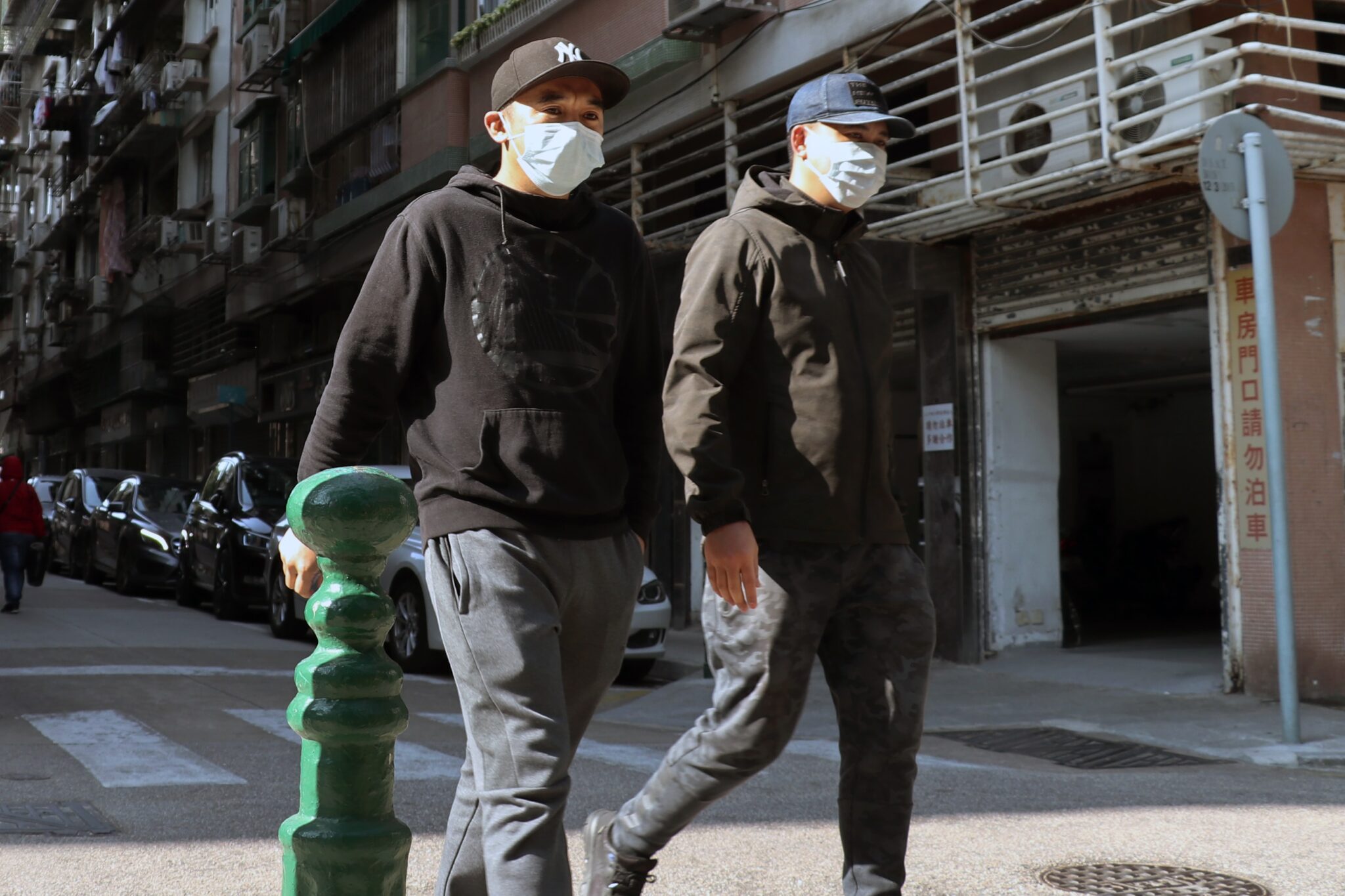 Dois indivíduos vestindo capuzes pretos, bonés de beisebol e máscaras faciais caminham lado a lado por uma rua da cidade de El Salvador. Eles passam por uma fileira de carros estacionados e um prédio com grades verticais de metal e uma porta de garagem parcialmente aberta.