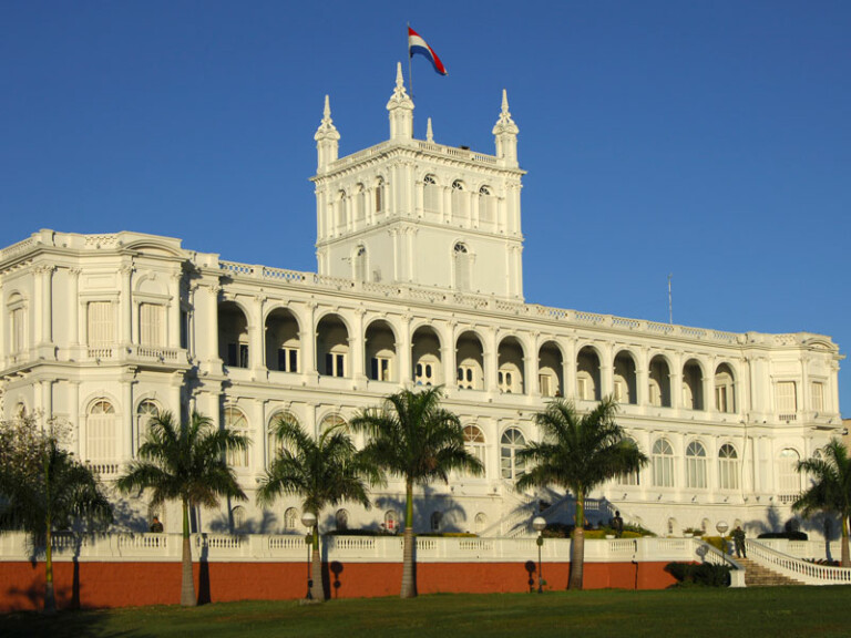 The image shows a large white neoclassical building with multiple arches and columns, set against a clear blue sky. A flag with red, white, and blue stripes flies on top. Palm trees line the front, and a grassy lawn in the foreground is part of the landscape, evoking a serene setting for discussing transfer pricing in Paraguay.