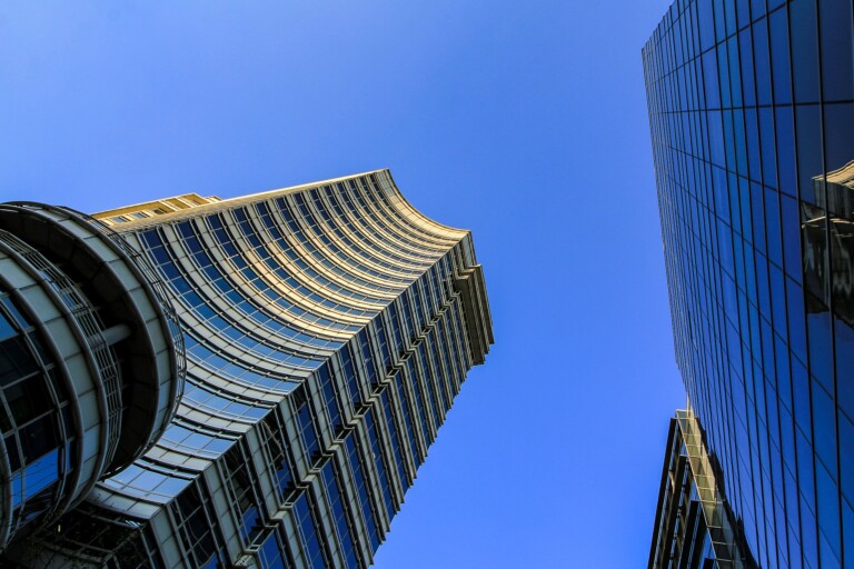 Upward view of modern skyscrapers with glass exteriors against a clear blue sky. The buildings display reflective surfaces and curved architectural elements, converging towards the top of the image, reminiscent of designs that could inspire new branches in Bolivia's urban landscape.