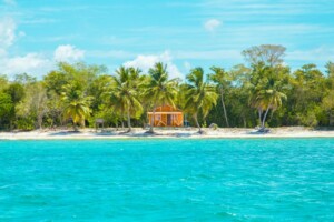 Una pequeña casa naranja con balcón se encuentra en una playa de arena, rodeada de altas palmeras y exuberante vegetación. El agua clara color turquesa del océano está en primer plano, con un cielo azul brillante en lo alto: un refugio perfecto para un abogado corporativo en República Dominicana.