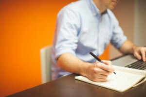 A person wearing a light blue shirt is sitting at a wooden table, writing in a notebook with a pen in their right hand and typing on a laptop with their left, likely planning to abrir uma filial na Bolívia. The background features an orange wall. The person's face is not visible.