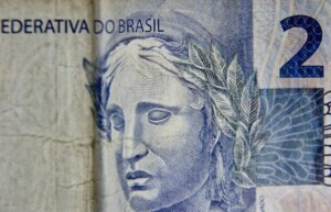 Primer plano de un billete de 2 reales brasileños. El lado izquierdo muestra parte del texto "FEDERATIVA DO BRASIL" y el lado derecho muestra el numeral "2". Una imagen intrincada de un rostro escultórico adornado con hojas de laurel simboliza la transferencia de precios en Brasil.