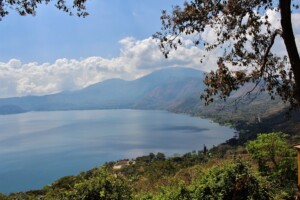 A vast lake surrounded by greenery and trees in the foreground, with mountains and cloudy skies in the background. The water is calm and blue under the daylight, seen from an elevated vantage point, capturing the expansive natural beauty similar to discovering back office services in El Salvador.