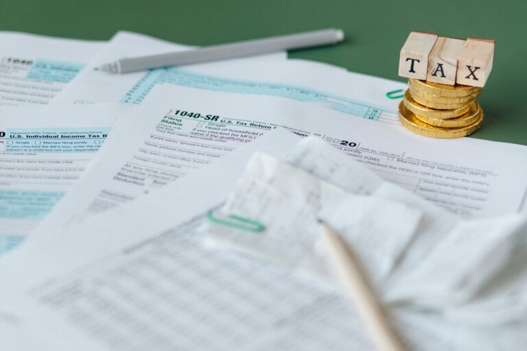 A collection of tax documents, including a 1040-SR form, is spread out on a table. In the background, gold coins are stacked with letter blocks spelling "TAX" on top. A pen and some receipts are also visible on the paperwork, emphasizing the importance of planejamento tributário corporativo.