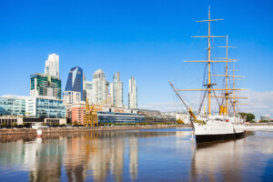 A sailboat with three tall masts is docked in a calm body of water. Modern high-rise buildings and a blue sky dominate the background, reminiscent of SAS in Argentina. The buildings and the boat are mirrored in the reflective water surface.