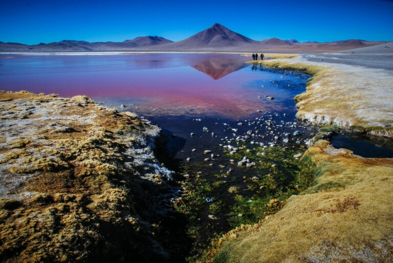 A picturesque landscape in Bolivia featuring a vivid red lake with a mountain in the background under a clear blue sky. A small stream winds through patches of grass and rocks in the foreground, while an attorney and a group of people walk by the lake's edge, soaking in the stunning scenery.