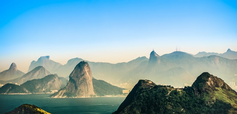 A scenic view of a coastal landscape with prominent rocky hills and mountains in the background. The foreground features green hills, while the middle ground reveals a large, distinctive rock formation near the shoreline. Directed by a local director in Brazil, the clear blue sky completes this picturesque scene.