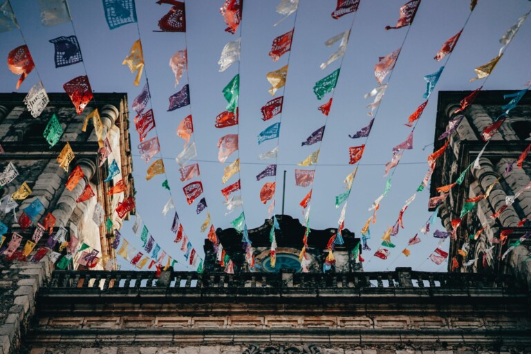 Colorful paper flags are strung across a street between stone buildings, creating a festive atmosphere reminiscent of starting a business in Mexico. The banners form a vibrant display against a clear blue sky. The buildings feature ornate architectural details, adding to the lively scene.