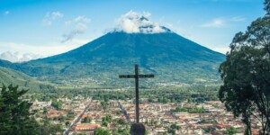 Una gran cruz se encuentra en lo alto de una colina que domina la ciudad de Antigua, Guatemala. Al fondo, se eleva prominentemente el volcán Volcán de Agua, con su cima parcialmente cubierta por nubes. El cielo azul claro contrasta con el exuberante paisaje verde.