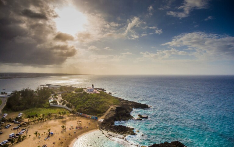 Aerial view of a coastal area in Costa Rica featuring a small sandy beach with people. There is a lighthouse on a rocky point extending into the ocean. The sun peeks through clouds on the left side, casting a warm glow over the scene. The ocean is calm with gentle waves, showcasing nature's due diligence.