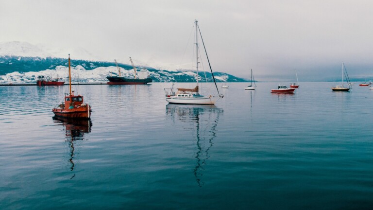 A calm body of water with several boats floating, including a sailboat in the foreground. Snow-covered mountains are visible in the background under a cloudy sky. The water reflects the boats and surrounding scenery, much like how clear invoicing requirements for a foreign company in Chile provide transparency.