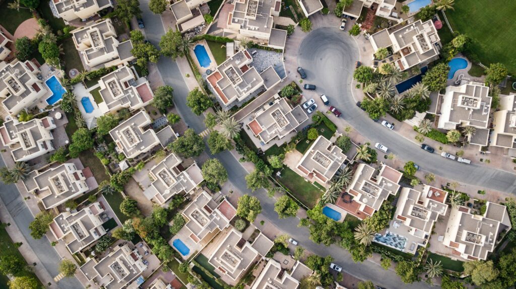 Aerial view of a residential neighborhood with multiple houses, some with pools. The area has curving roads and lush greenery. Vehicles are parked along the streets and in driveways. The houses are uniformly designed with beige rooftops, offering a picturesque setting to buy property in Santiago.