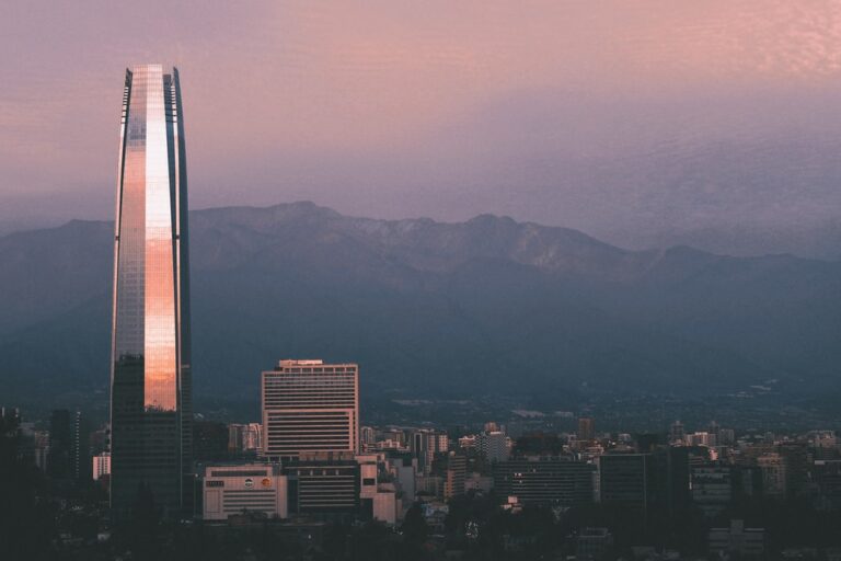 A city skyline at dusk with a mix of tall buildings and smaller structures. A prominent, illuminated skyscraper is seen on the left, with its reflective surface catching the light. Mountains are visible in the background under a pinkish sky, highlighting why so many choose to buy property in Santiago.