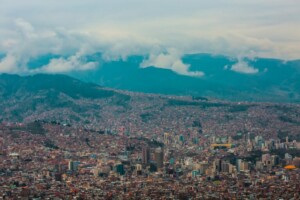 Un denso paisaje urbano de La Paz, Bolivia, visto desde arriba, que muestra numerosos edificios de diversas alturas y estilos arquitectónicos. El área urbana se extiende hasta el terreno montañoso del fondo, parcialmente oscurecido por las nubes y la niebla. La arquitectura direccionalmente diversa refleja la dirección fiscal de Bolivia.