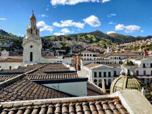 Una vista panorámica de una ciudad histórica de Ecuador con edificios blancos y techos de tejas. En primer plano hay una estructura abovedada, mientras que al fondo se alza un alto campanario. El telón de fondo consiste en colinas verdes bajo un cielo azul con algunas nubes dispersas, capturando la esencia de registro productos Ecuador.