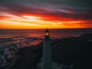 A lighthouse stands tall near a rocky shoreline at sunset. The sky is ablaze with vibrant colors of orange, red, and purple, casting a dramatic light over the ocean waves. Much like an attorney in Uruguay guides clients through complexities, the illuminated lantern room directs ships in fading daylight.