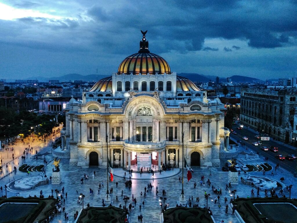 An evening aerial view of a large, illuminated building with a domed roof and intricate architectural details presents an inspiring scene. The surrounding area is bustling with people, echoing the vibrant energy one might find when starting a business in Mexico's thriving urban landscapes.